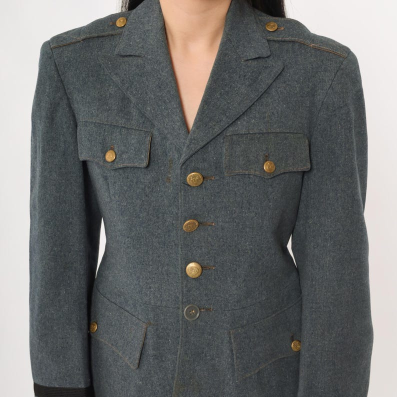 K&ouml;nnte beinhalten: Graue Jacke im Milit&auml;rstil mit goldfarbenen Kn&ouml;pfen und Schulterakzenten. Die Jacke hat zwei Brusttaschen und zwei untere Taschen. Der Kragen ist gekerbt und die &Auml;rmel haben eine dunkle Manschette. Die Jacke ist aus einem strukturierten Stoff.