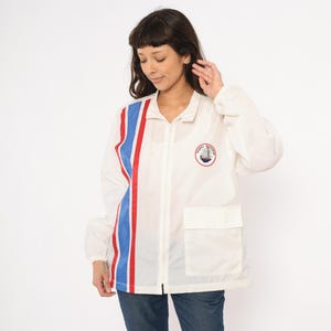 May include: White zip-up jacket with a red and blue stripe down the left side. The jacket has a small pocket and a circular emblem with a sailboat. The jacket is made of a lightweight material.