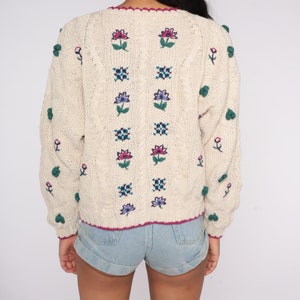 May include: A cream-colored knitted sweater with a floral design and pom-poms on the sleeves. The sweater has a V-neck and a cable knit pattern.