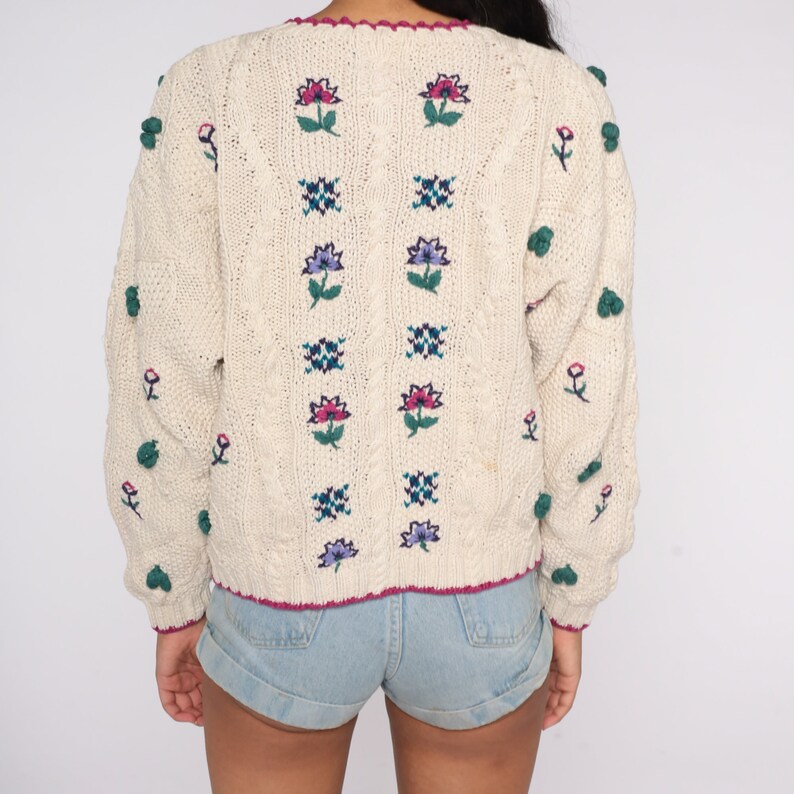 May include: A cream-colored knitted sweater with a floral design and pom-poms on the sleeves. The sweater has a V-neck and a cable knit pattern.