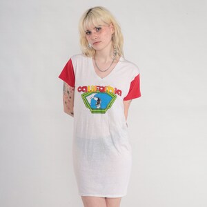 K&ouml;nnte beinhalten: Wei&szlig;es T-Shirt-Kleid mit roten &Auml;rmeln und einem Grafikdruck eines Surfers vor einem blauen und gr&uuml;nen Kalifornien-Logo. Der Text "california" ist in Gelb und Orange.