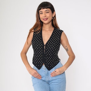 May include: Black vest with white polka dots. The sleeveless vest has a V-neck and button closure. The vest is worn with light blue jeans.