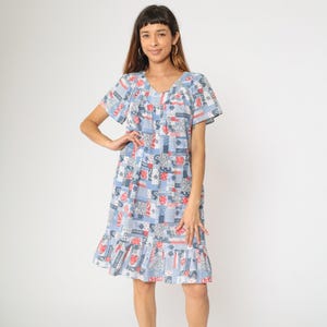 May include: A knee-length, short-sleeved dress with a V-neckline. The dress features a patchwork pattern in shades of blue, red, and white. The fabric has a ruffle at the hemline. The dress is worn by a person.
