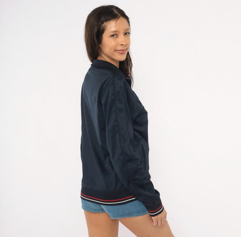 May include: A navy blue track jacket featuring a high collar, long sleeves, and a front zipper closure. The jacket includes ribbed cuffs and a ribbed hem, both detailed with thin white and red horizontal stripes. The fabric has a smooth finish.