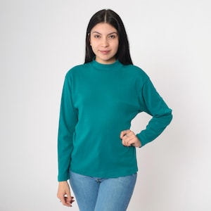 Pode incluir: Camisa de manga comprida verde-azulado com gola alta. A camisa &eacute; feita de um material macio e &eacute; combinada com jeans azuis. A modelo est&aacute; em frente a um fundo branco.