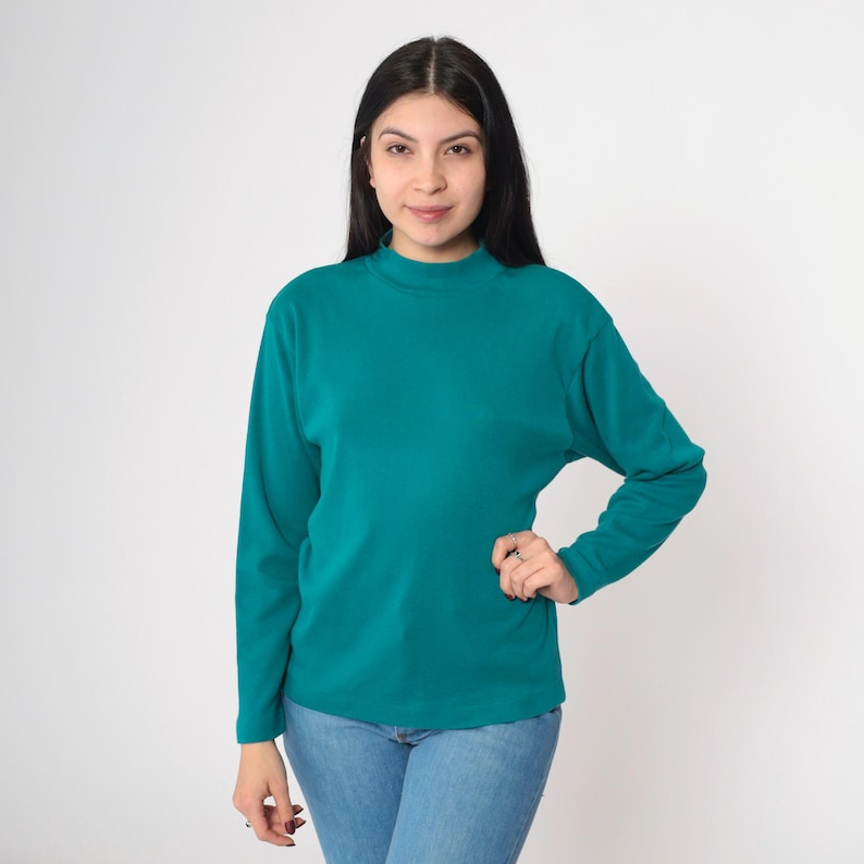 Pode incluir: Camisa de manga comprida verde-azulado com gola alta. A camisa &eacute; feita de um material macio e &eacute; combinada com jeans azuis. A modelo est&aacute; em frente a um fundo branco.