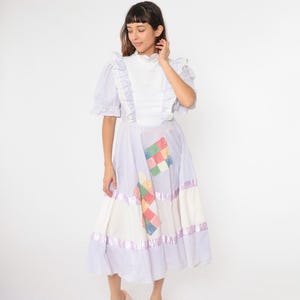 Può includere: Un abito a righe bianche e lilla con un colletto bianco a balze e dettagli in tessuto patchwork. L'abito ha una gonna a pieghe e un corpetto aderente.