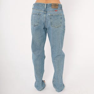 Peut inclure: Jean bleu clair en denim avec une conception classique &agrave; cinq poches. Le jean a une coupe droite et un patch en cuir marron sur la ceinture arri&egrave;re. L'&eacute;tiquette de la marque Wrangler est visible.