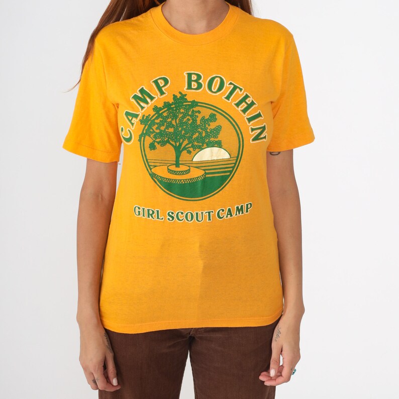May include: Mustard yellow t-shirt with a green and white graphic. The graphic features a tree, a setting sun, and the text "CAMP BOTHIN GIRL SCOUT CAMP" in a circular design. The shirt has short sleeves and a crew neck.