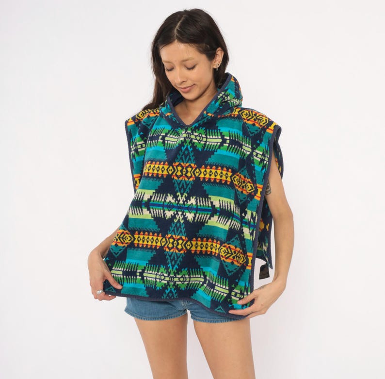 May include: A hooded poncho featuring a vibrant geometric pattern in shades of navy blue, teal, lime green, yellow, and orange. The garment is constructed from a soft fleece material and includes a hood, wide arm openings, and dark blue trim.