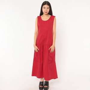 May include: A sleeveless, ankle-length red dress featuring a round neckline and a dropped waistline. The garment includes two large, rectangular patch pockets positioned on the front of the skirt section, which has a gathered, full-length silhouette.