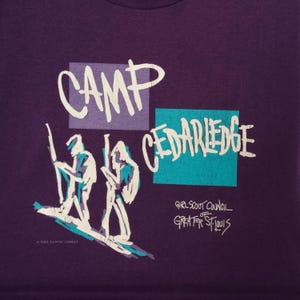 May include: Purple t-shirt with a graphic design featuring the words "CAMP CEDARLEDGE" in white, along with stylized figures and text that reads "GIRL SCOUT COUNCIL GREATER ST. LOUIS". The design includes a copyright notice.