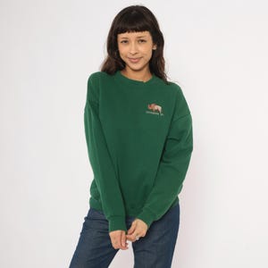 May include: A green sweatshirt with a crew neck and long sleeves. The sweatshirt features an embroidered image of a bison and the text "CHEYENNE, WY" on the upper left chest. The person is wearing blue jeans.