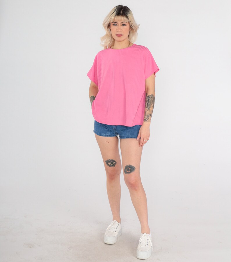May include: A pink short-sleeved shirt with a ribbed texture. The model is wearing denim shorts and white sneakers.