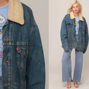 Puede incluir: Una chaqueta de mezclilla azul con cuello de sherpa color crema, cierre de botones met&aacute;licos y dos bolsillos en el pecho. Debajo, una camiseta azul claro con un gr&aacute;fico de un gato y el texto NAP ATTACK! junto a pantalones vaqueros anchos.
