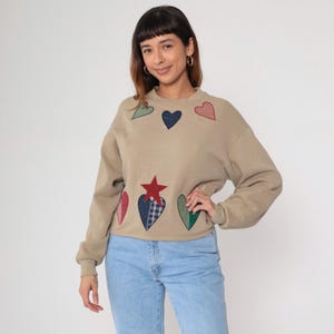 May include: Beige sweatshirt featuring appliqued hearts and a star in various colours. The hearts are red, green, and blue. The star is red and blue. The sweatshirt has a crew neck and long sleeves. The model is wearing blue jeans.