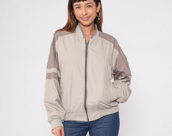 May include: A beige bomber jacket with brown shoulder panels and sleeve stripes. The jacket has a zip-up front, a ribbed collar, and ribbed cuffs. The model is wearing blue jeans.