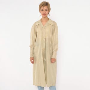 May include: A long-sleeved, knee-length raincoat in a solid beige color. The coat features a pointed collar, a button-down front closure with matching beige buttons, and two vertical pockets on the front. The material has a smooth, semi-sheer finish.