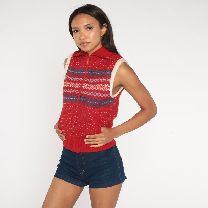 May include: A red sleeveless knitted vest with a white collar and a zip closure. The vest has a geometric pattern in blue, white, and pink.