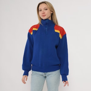 May include: A blue sweater with a high collar and a quarter-zip closure. The shoulders feature red and yellow color blocks. The sweater has long sleeves and a ribbed hem and cuffs. The model is wearing light blue jeans.