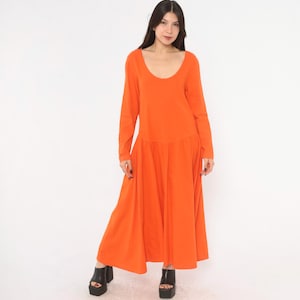 May include: A long-sleeved, floor-length dress in a solid bright orange colour. The garment features a wide scoop neckline and a fitted bodice that transitions into a full, flowing skirt with a dropped waistline, creating a simple silhouette.
