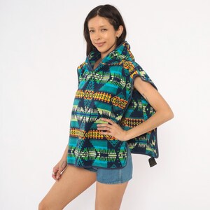 May include: A hooded poncho featuring a vibrant geometric pattern in shades of teal, navy blue, yellow, and orange. The garment is constructed from a soft fleece material and includes a hood, wide arm openings, and a hem that falls at the hip.