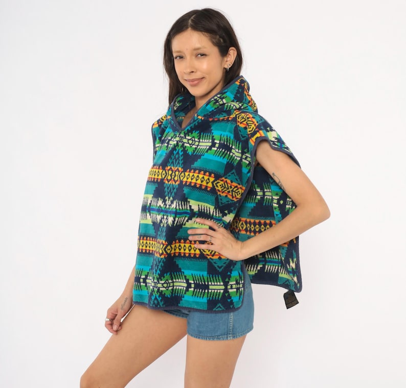 May include: A hooded poncho featuring a vibrant geometric pattern in shades of teal, navy blue, yellow, and orange. The garment is constructed from a soft fleece material and includes a hood, wide arm openings, and a hem that falls at the hip.