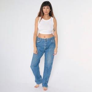 May include: A woman wearing a white vest top and blue denim jeans. The jeans have a straight leg fit and a high waist.