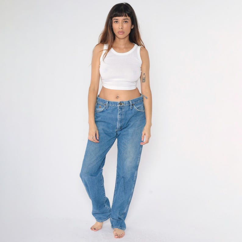 May include: A woman wearing a white vest top and blue denim jeans. The jeans have a straight leg fit and a high waist.