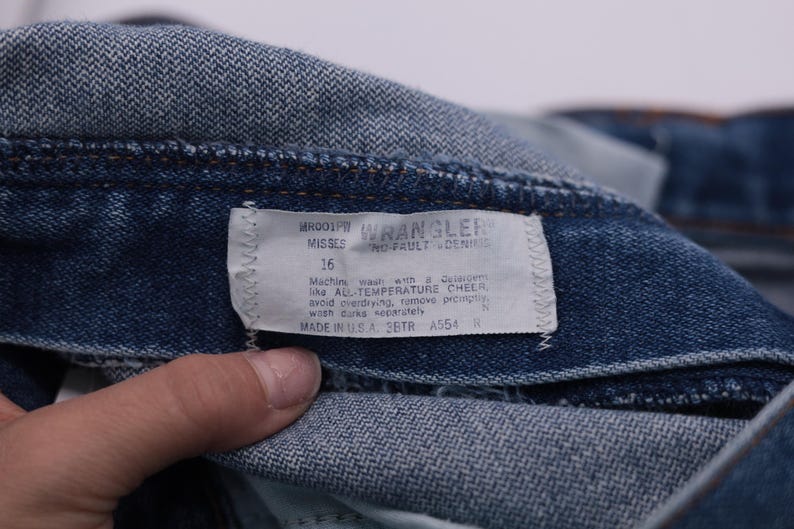 May include: Close-up of a blue denim Wrangler jeans label with the text "ROOP WRANGLER MISSES 16 WO-PAULT DENIM" and care instructions. Machine wash with All-Temperature Cheer, avoid overdrying, remove promptly, wash dark separately. Made in USA SBTR A554.