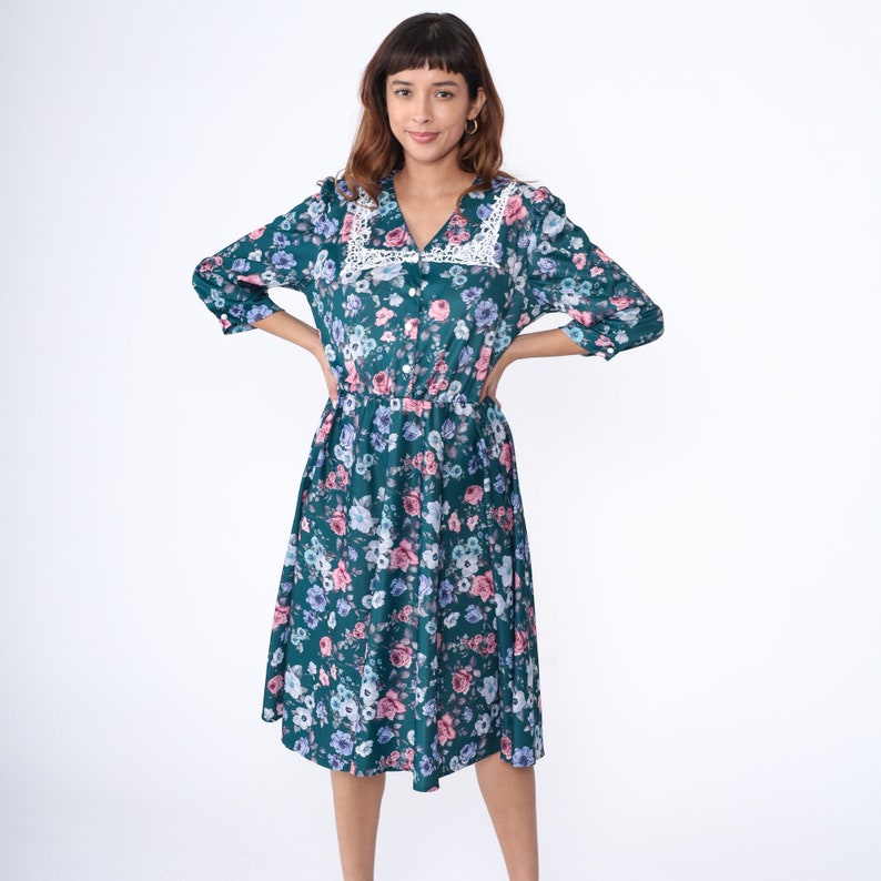 K&ouml;nnte beinhalten: Ein blaugr&uuml;nes Blumenkleid mit einem wei&szlig;en Spitzenkragen. Das Kleid hat einen Knopfleisten vorne und eine geraffte Taille.
