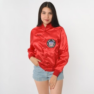 May include: A red satin bomber jacket with a button-up front and a ribbed collar and cuffs. The jacket features a patch on the chest with the words "United States Karate Association". The jacket is worn with denim shorts.