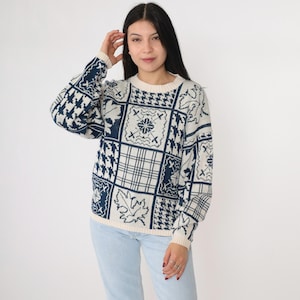 Puede incluir: Suéter color crema con patrones geométricos azul marino. El suéter presenta cuello redondo y mangas largas. El diseño incluye cuadrados con cuadros, hojas y otros motivos decorativos. El suéter se usa con jeans azul claro.