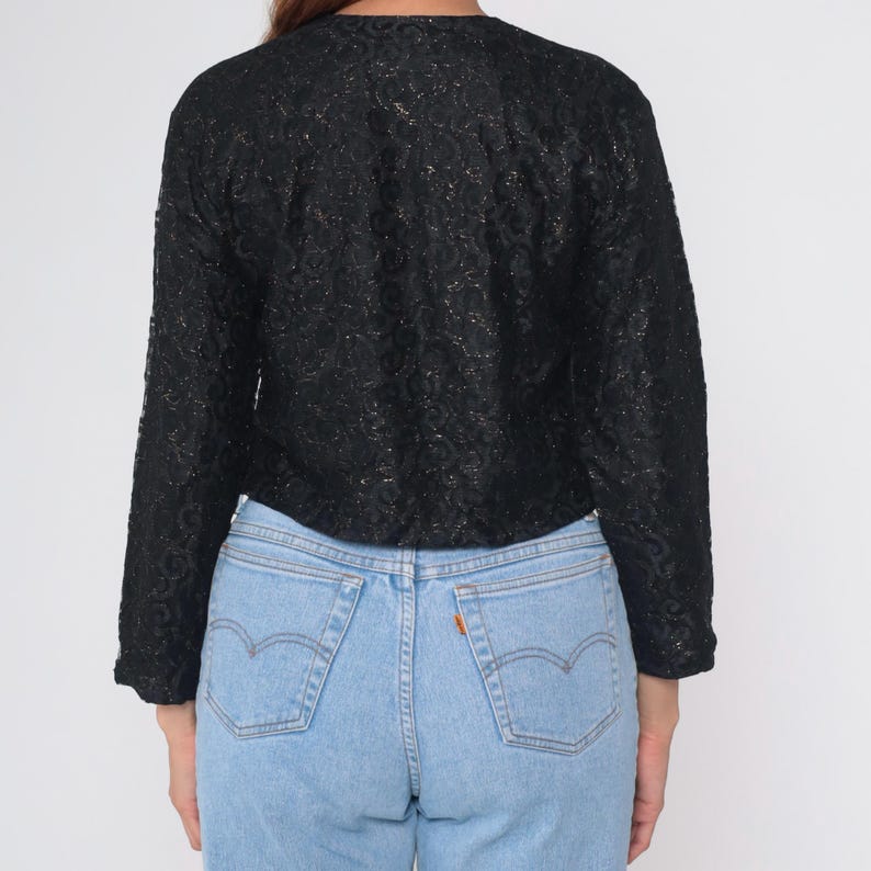 Puede incluir: Una blusa negra de manga larga con un patr&oacute;n floral texturizado. Se combina con vaqueros de mezclilla azul claro. La vista trasera muestra el dise&ntilde;o y el ajuste de la ropa, adecuada para uso casual o semi-formal.