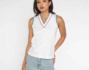 Può includere: Top bianco senza maniche con scollo a V. Lo scollo è rifinito con un bordo blu acqua e una striscia interna rossa. Il top è abbinato a jeans di denim blu chiaro. La modella è davanti a uno sfondo bianco.