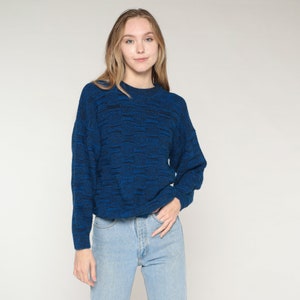 Peut inclure: Un pull bleu en maille avec un col rond. Le pull présente un motif texturé avec des fils bleu foncé et noirs. Le modèle porte un jean bleu clair. Le fond est blanc uni.