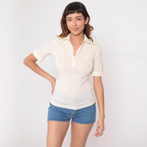 Puede incluir: Polo crema con mangas cortas y cuello con cremallera. La camisa tiene un pequeño emblema bordado en el pecho. Se usa con pantalones cortos de mezclilla.