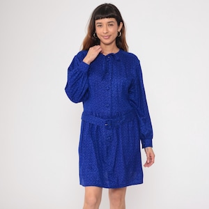 May include: A vintage cobalt blue dress with a subtle patterned design. The dress features long sleeves, a button-down front, and a matching belt with a buckle. The dress has a collared neckline and a knee-length skirt.