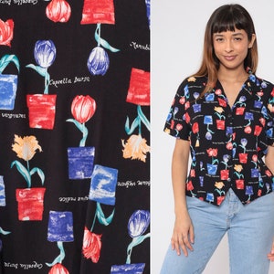 Op de afbeelding: Zwart shirt met korte mouwen en een bloemenpatroon. Het patroon bevat kleurrijke tulpen in potten met botanische labels. Het shirt heeft een V-hals en wordt gedragen met een lichtblauwe jeans.
