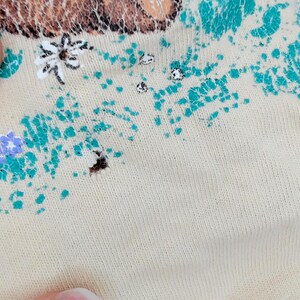 May include: Close-up of a beige fabric with a floral design. The design includes teal, purple, and white floral elements. The fabric appears to be a knit material.