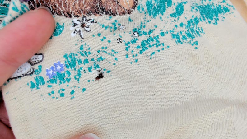 May include: Close-up of a beige fabric with a floral design. The design includes teal, purple, and white floral elements. The fabric appears to be a knit material.