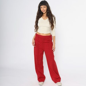 May include: A woman wearing a cream-colored, long-sleeved V-neck shirt and red wide-leg pants. The pants are a solid color and have a relaxed fit. The woman is wearing white sneakers.