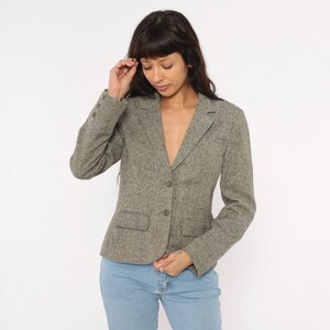 May include: A gray tweed blazer with a notched lapel, two front pockets, and button closures. The blazer is worn over a light-colored top and paired with blue jeans. The jacket has a classic, tailored fit.