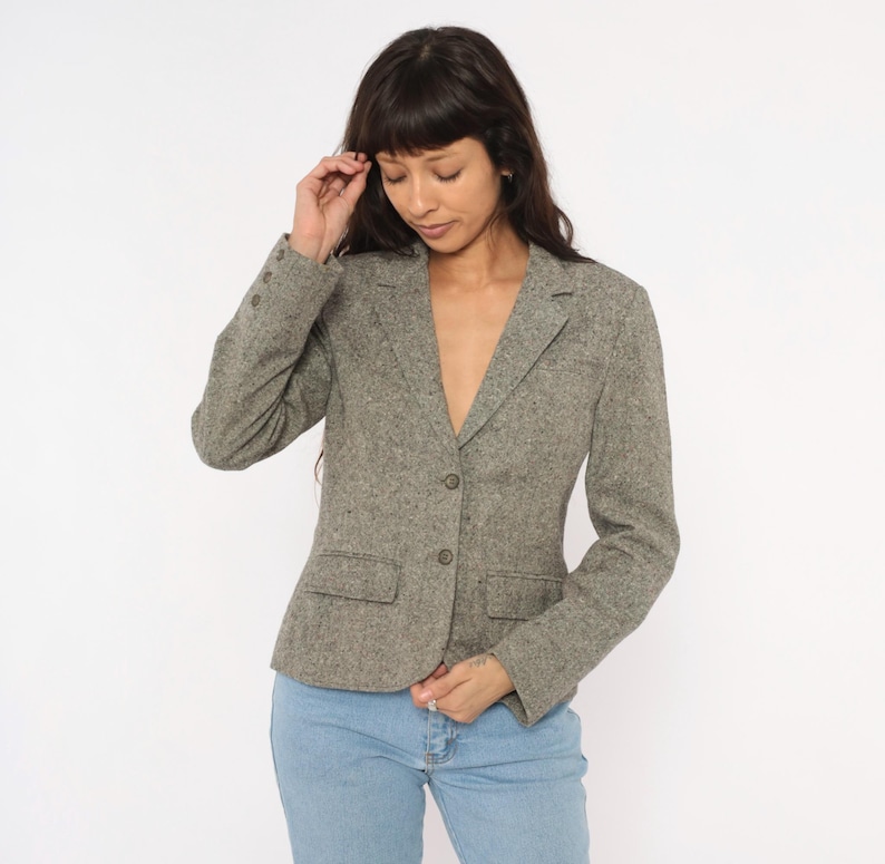 May include: A gray tweed blazer with a notched lapel, two front pockets, and button closures. The blazer is worn over a light-colored top and paired with blue jeans. The jacket has a classic, tailored fit.