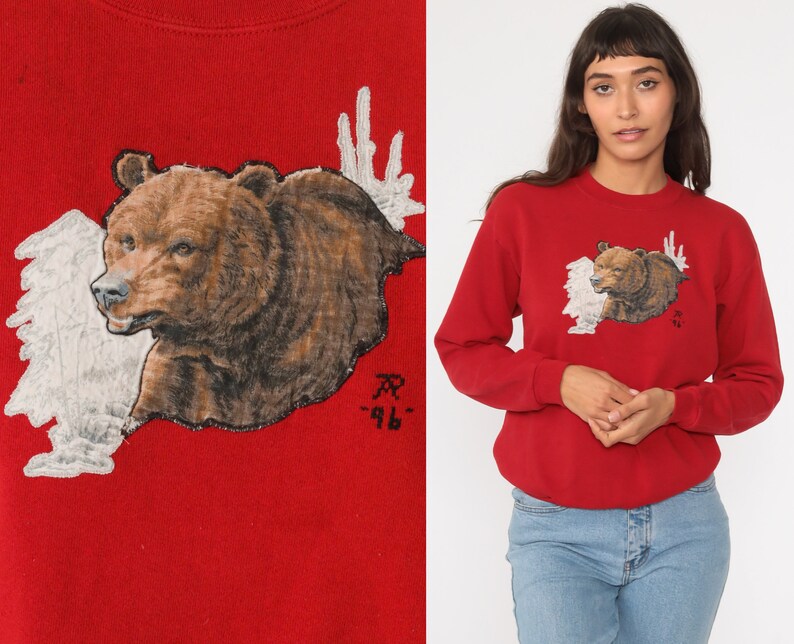 May include: Red sweatshirt with a brown bear and white antlers graphic. The bear is facing left and the antlers are behind it. The text "AR -96" is printed below the bear.