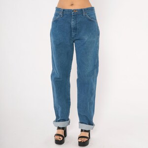 May include: Blue denim jeans with a classic straight leg fit. The jeans have a button and zipper closure, front and back pockets, and are cuffed at the ankles. The model is wearing black platform sandals.