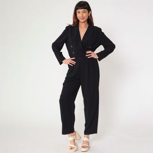 May include: A black jumpsuit with a double-breasted blazer-style top and wide-leg pants. The blazer features gold-colored buttons. The model is wearing cream-colored platform heels. The background is plain white.