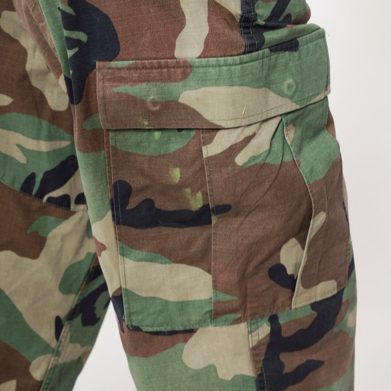 May include: Close-up of camouflage cargo pants. The pants feature a classic woodland camouflage pattern in shades of green, brown, and black. A pocket is visible on the side. The fabric appears to be durable and suitable for outdoor activities.