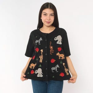 Op de afbeelding: Een zwart shirt met korte mouwen en knoopsluiting, voorzien van geborduurde safaridieren zoals olifanten, giraffen, zebra's, een aap en een tijger, afgewisseld met rode hibiscusbloemen en kleine groene grasaccenten op de voorzijde.