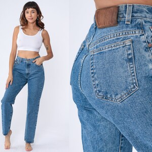 May include: Light blue denim jeans with a classic straight-leg fit. The jeans feature a high waist, front pockets, and a brown leather patch on the back. The model is wearing a white vest top. The jeans are made of a durable denim material.